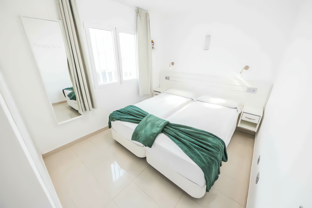 Helles Schlafzimmer mit einem Doppelbett, einer grünen Decke und einem Spiegel an der Wand.