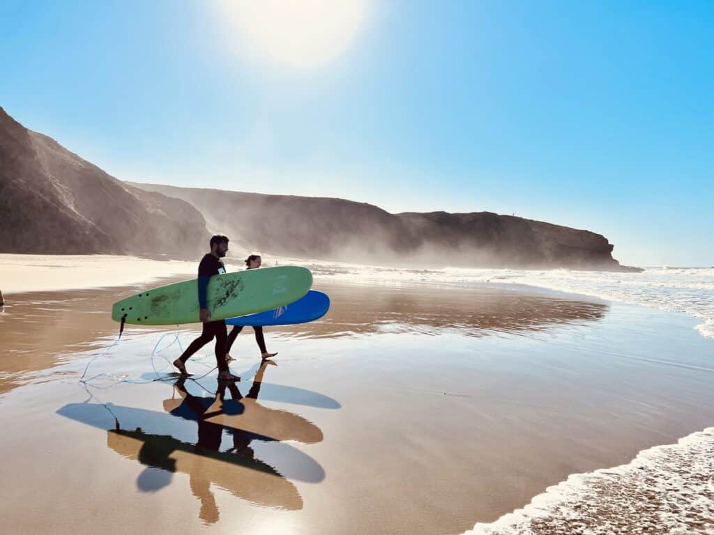 Two surfers are carrying green and blue surfboards along the beach in sunny weather.