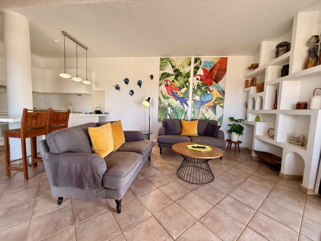 Wohnzimmer mit Sofa und Wandmalerei Wohnzimmer mit grauem Sofa, gelben Kissen und einer bunten Wandmalerei von Papageien.