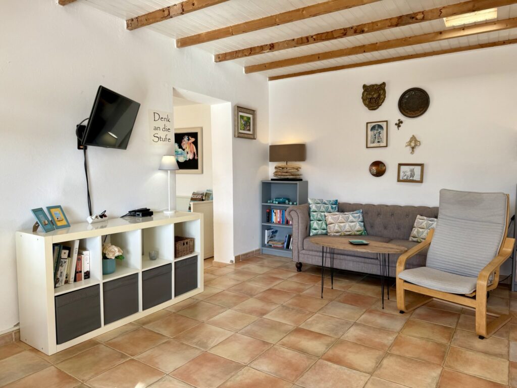Ein Wohnzimmer mit einem grauen Sofa, einem Fernseher an der Wand und einem Holzstuhl.