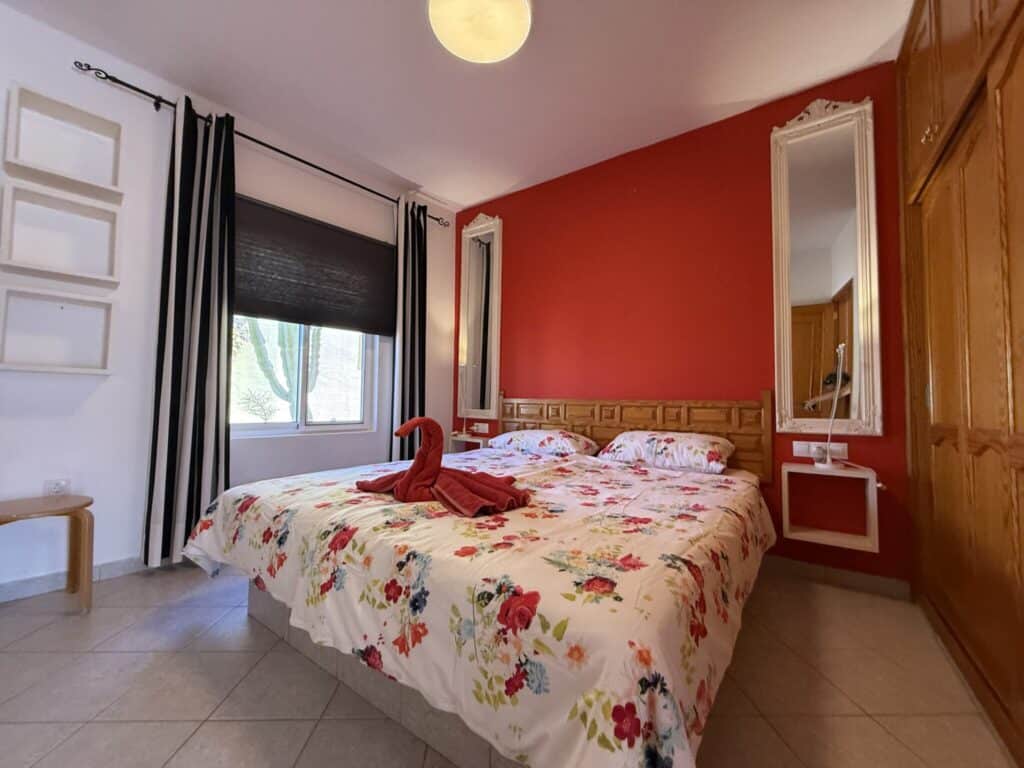 Schlafzimmer mit einem Doppelbett, roter Wand und Blumenbettwäsche in einem hellen Raum.